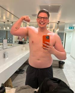A shirtless man flexing his bicep while taking a selfie in a gym locker room.