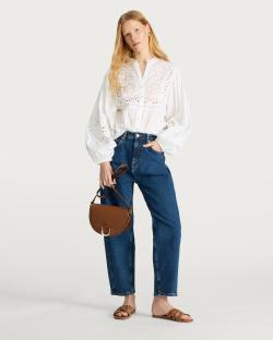 Woman in eyelet blouse, jeans, and brown sandals carrying a brown bag.