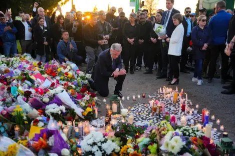 A man kneels among a large display of flowers and lit candles at a vigil.