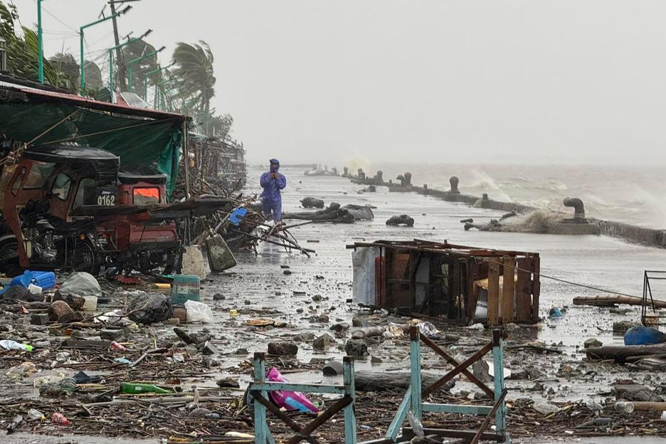 A man in a raincoat standing on a debris-strewn waterfront road with large waves crashing in the background due to Super Typhoon Ragasa.