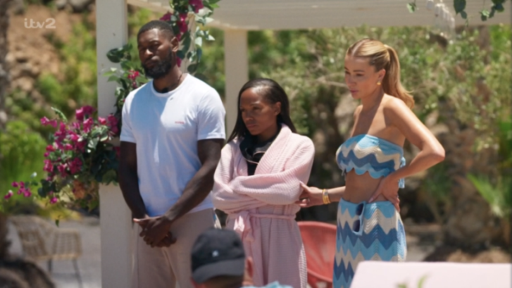 Three contestants of the reality show Love Island UK, two women and one man, stand outside, looking intently at someone off-camera.