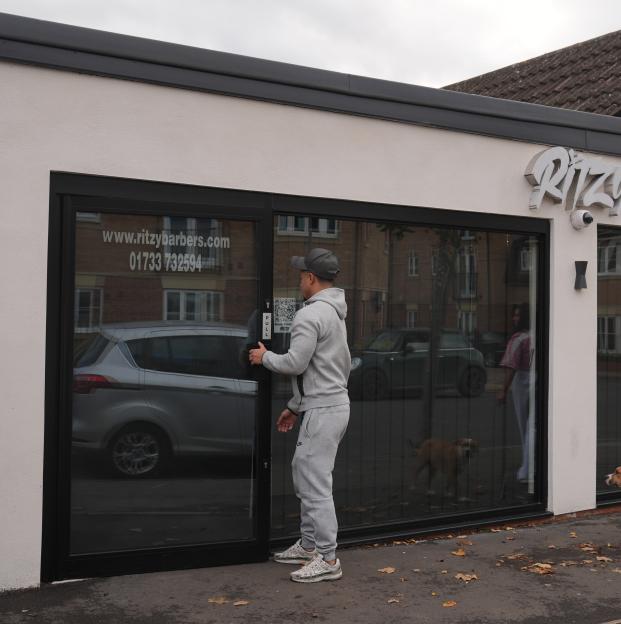 A man opening the door to the Ritzy Barbers in Fletton, with reflections of cars, a dog, and a person in the window.