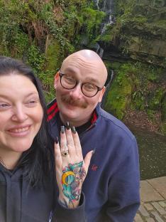 Couple posing for a selfie in front of a waterfall. The woman shows off her engagement ring.