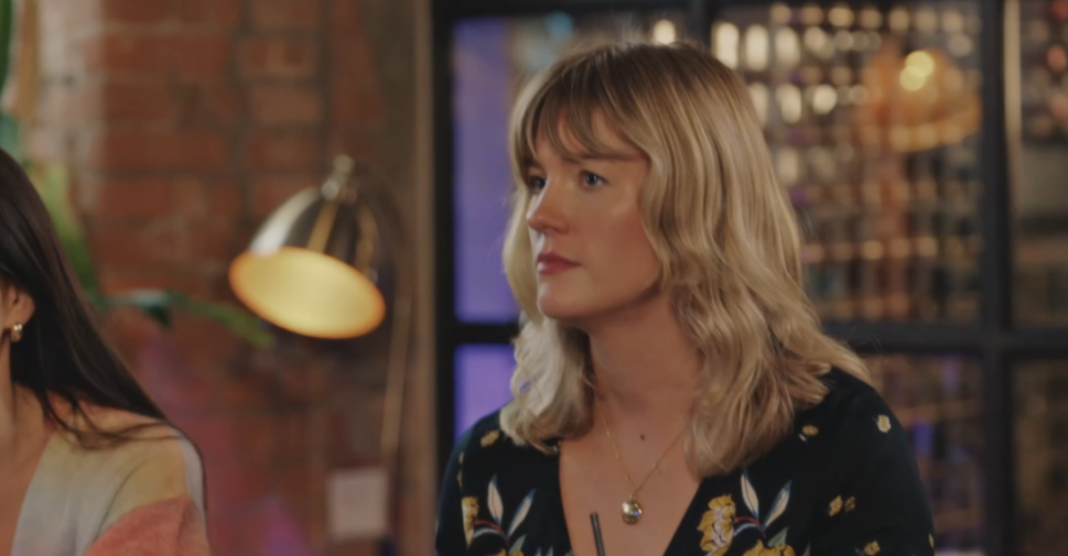A woman with blonde hair and a floral top listens intently in a dimly lit bar.