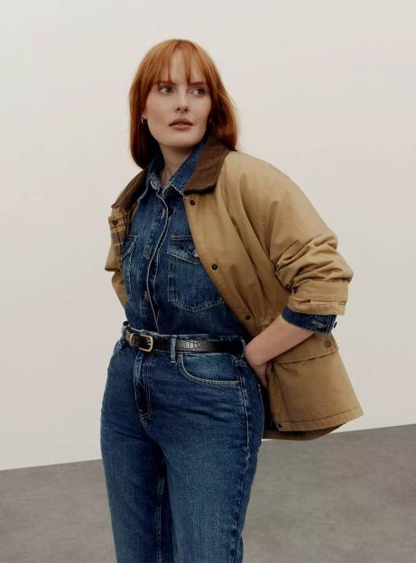 A model wearing a tan jacket with a corduroy collar over a denim shirt, dark-wash jeans, and a black belt.