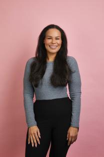 Smiling woman with long dark hair wearing a gray long-sleeve shirt and black pants.