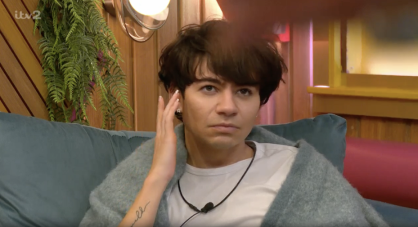 A person with dark messy hair, wearing a white t-shirt and light blue fuzzy blanket, sits on a blue couch with a serious expression and one hand near their ear.