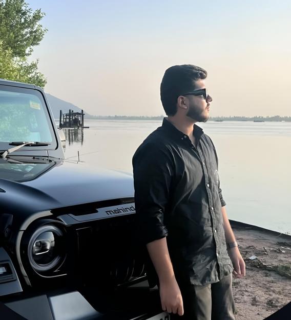 A young man in sunglasses stands beside a black Mahindra SUV, overlooking a calm body of water.