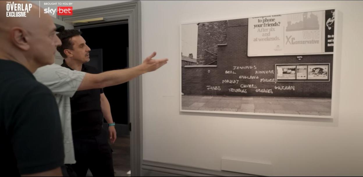 Photo of two men looking at a black and white photo of a brick wall with graffiti of soccer players' names.