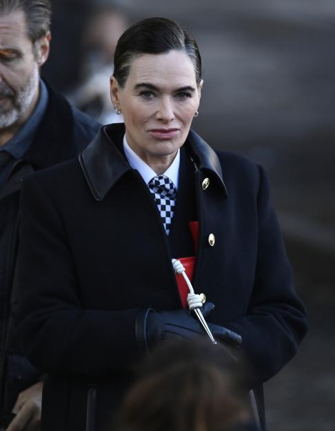 Actress Lena Headey in character with a slicked-back hairstyle, black coat, and checkerboard tie, on a film set with another actor partially visible.