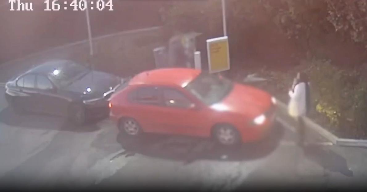 Security camera footage of a red car pulling up to a gas pump, behind a black car, with a person standing next to the red car.