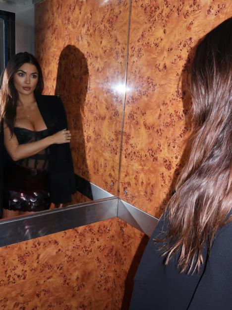 India Reynolds wearing a black top and jacket, looking at her reflection in a mirror.