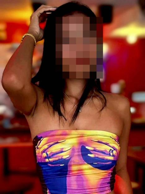 A woman with her face pixelated, wearing a colorful strapless top with a graphic of hands cupping breasts, and a gold bracelet.
