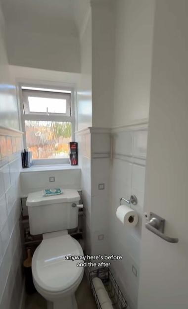 A toilet with white walls and tiles, a window, and a roll of toilet paper on the right.