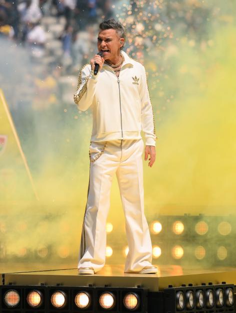 Robbie Williams performs on stage in a white and gold tracksuit with a microphone.