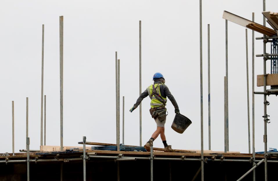 A construction worker in a hard hat, safety vest, and tool belt walking across scaffolding while carrying a bucket.