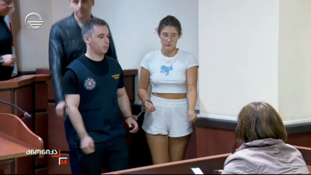 A woman in white shorts and a t-shirt, handcuffed, stands in a courtroom next to a guard.