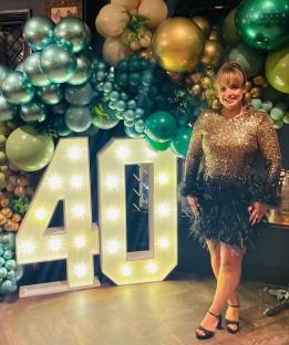 Woman in a gold and black feather dress standing in front of a 40th birthday balloon and light-up number display.