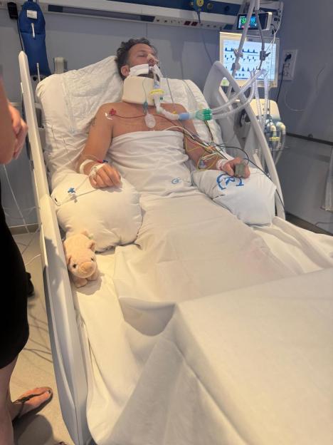 David Hayes, 37, in a hospital bed with a neck brace and medical tubes, with a stuffed pig on the bed.
