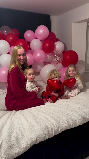 Woman with three young children on a bed with pink and red balloons.