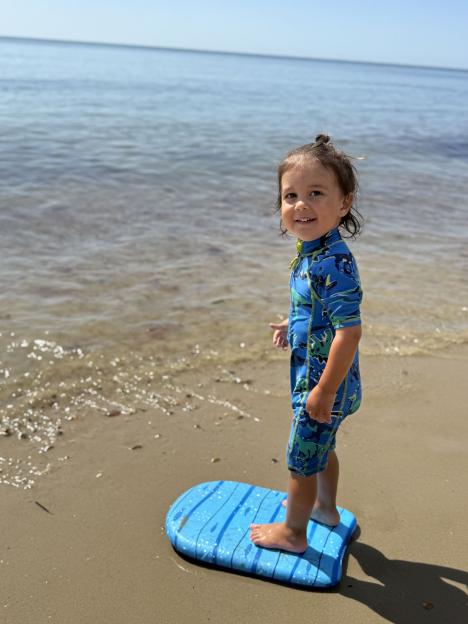 Toddler standing on a boogie board on a beach.