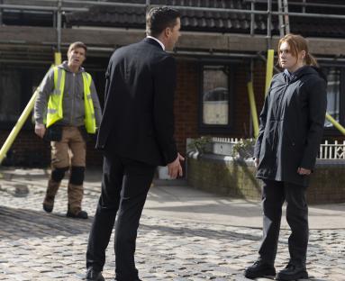 A man in a suit confronts a young woman, while a man in a high-visibility vest looks on.