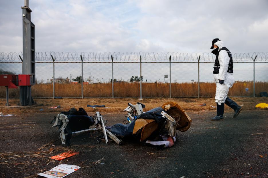 An investigator in a white suit and boots walks past wreckage from Jeju Air Flight 2216 at Muan International Airport.