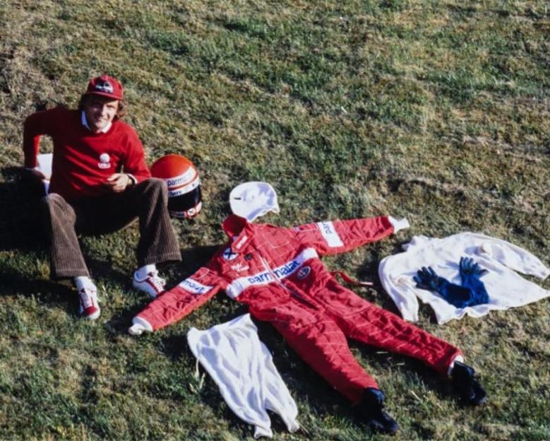 Photo of Niki Lauda sitting on the grass with his racing suit and helmet laid out beside him.