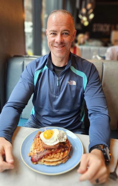 Dave Stewart sitting at a table with a stack of pancakes, beans, bacon, and a fried egg on a plate in front of him.