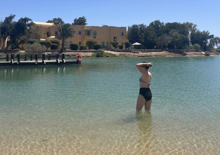 Person in a bathing suit standing in shallow water near a resort.