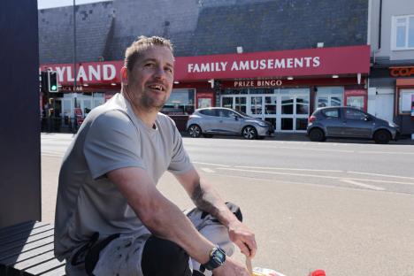 A man sits on a bench eating, with a family amusement arcade in the background.