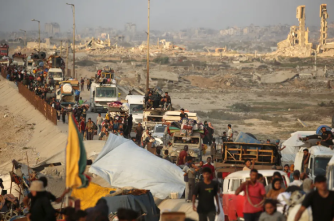 People and vehicles laden with belongings move along a road with rubble and damaged buildings in the background.