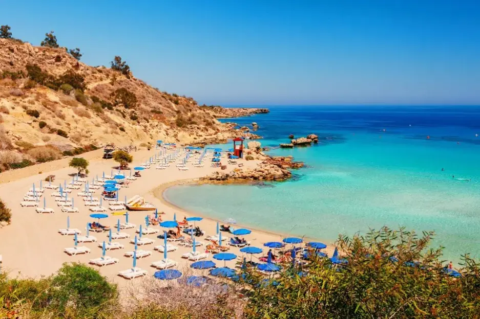 A beach with white lounge chairs and blue umbrellas on a sandy shore next to clear turquoise water, with a rocky hillside on the left.