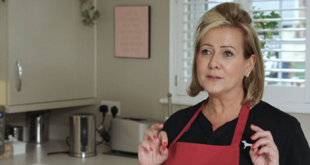 Woman in a red apron and black shirt speaking in a kitchen.