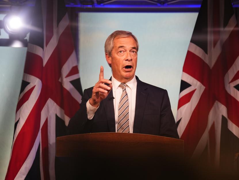 Reform UK leader Nigel Farage speaking at a podium with two Union Jack flags behind him.