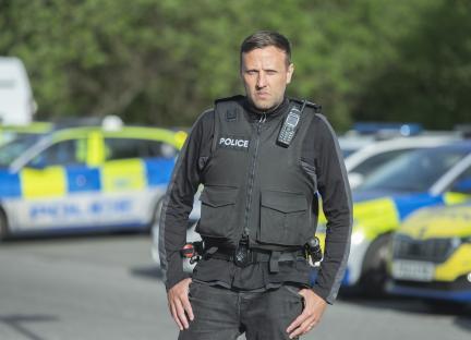 Police officer in tactical vest standing in front of police cars.