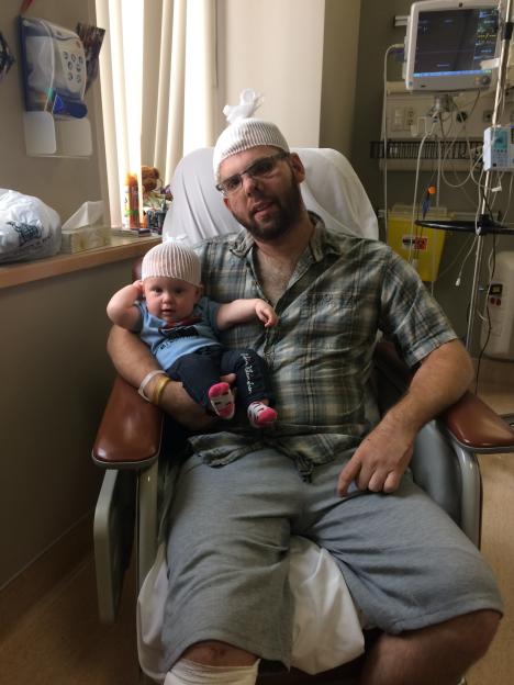 A man with head bandages holds a baby with head bandages in a hospital chair.
