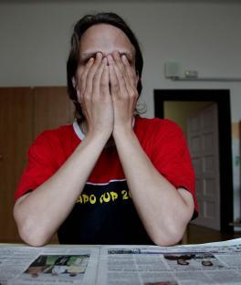 A person sits at a table with their face covered by their hands, leaning on a newspaper.