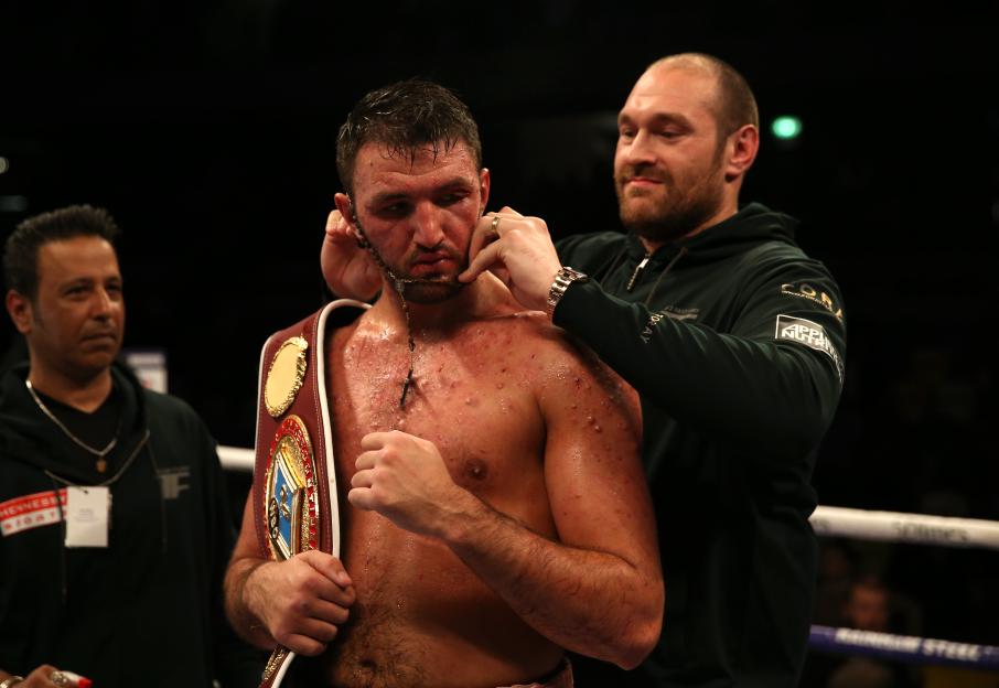 Tyson Fury places a rosary necklace on Hughie Fury, who is holding a WBO championship belt.