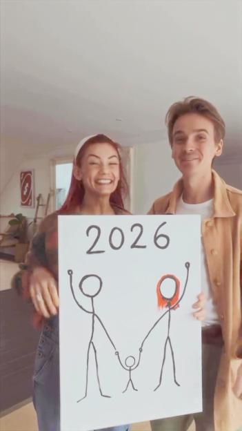 Dianne Buswell and Joe Sugg holding a sign announcing their pregnancy with a stick figure family drawing and "2026".