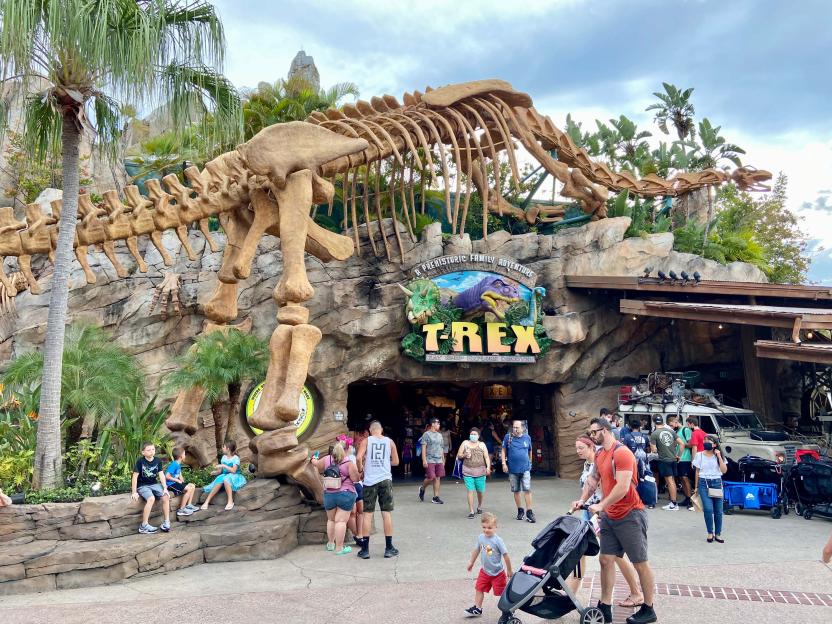 The entrance to T-Rex Cafe, a dinosaur-themed restaurant, with a large dinosaur skeleton arching over the entrance and people walking around.