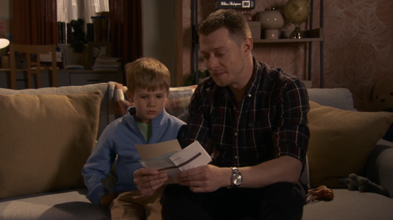 Screenshot of a man and a young boy sitting on a couch, looking at a piece of mail.