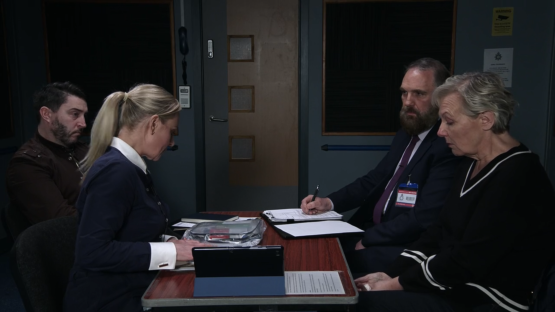 Screenshot of four people sitting around a table in an interview room, one person is writing.