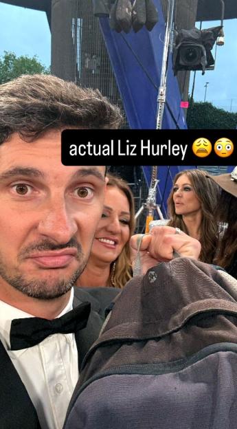 A man in a tuxedo poses for a selfie with Liz Hurley and other actors in the background.