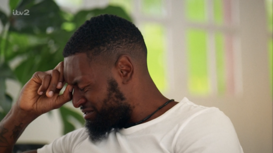 A man with short dark hair and a beard, wearing a white t-shirt and a dark necklace, grimaces with his eyes closed and his hand pressed to his forehead.
