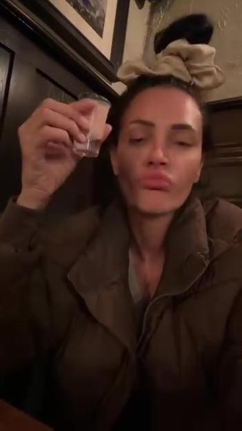 A woman in a dark jacket holds up a shot glass while pouting her lips at the camera.
