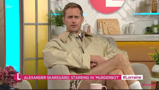 Alexander SkarsgÃ¥rd on the Lorraine show wearing a trench coat and shorts.