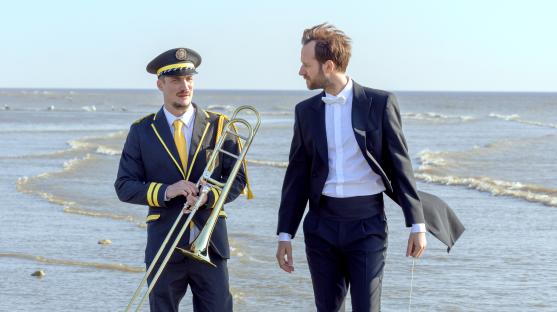 Film still of two men in formal wear on a beach; one plays a trombone.