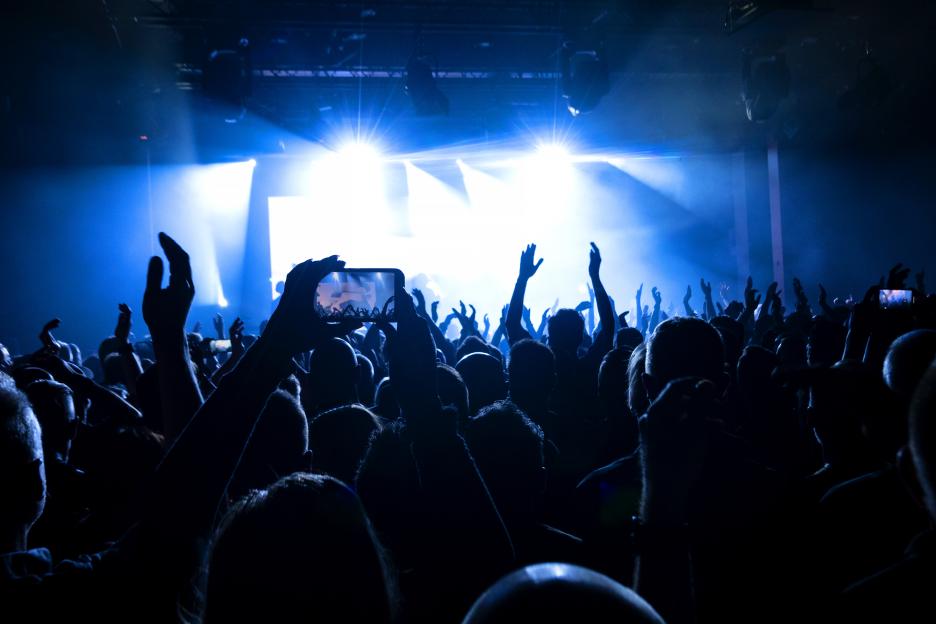 Silhouetted concert crowd with raised hands; some are recording the performance with phones.
