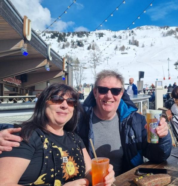 Patrick Howard with his wife Laura, smiling and holding drinks at an outdoor table with a snowy mountain in the background.